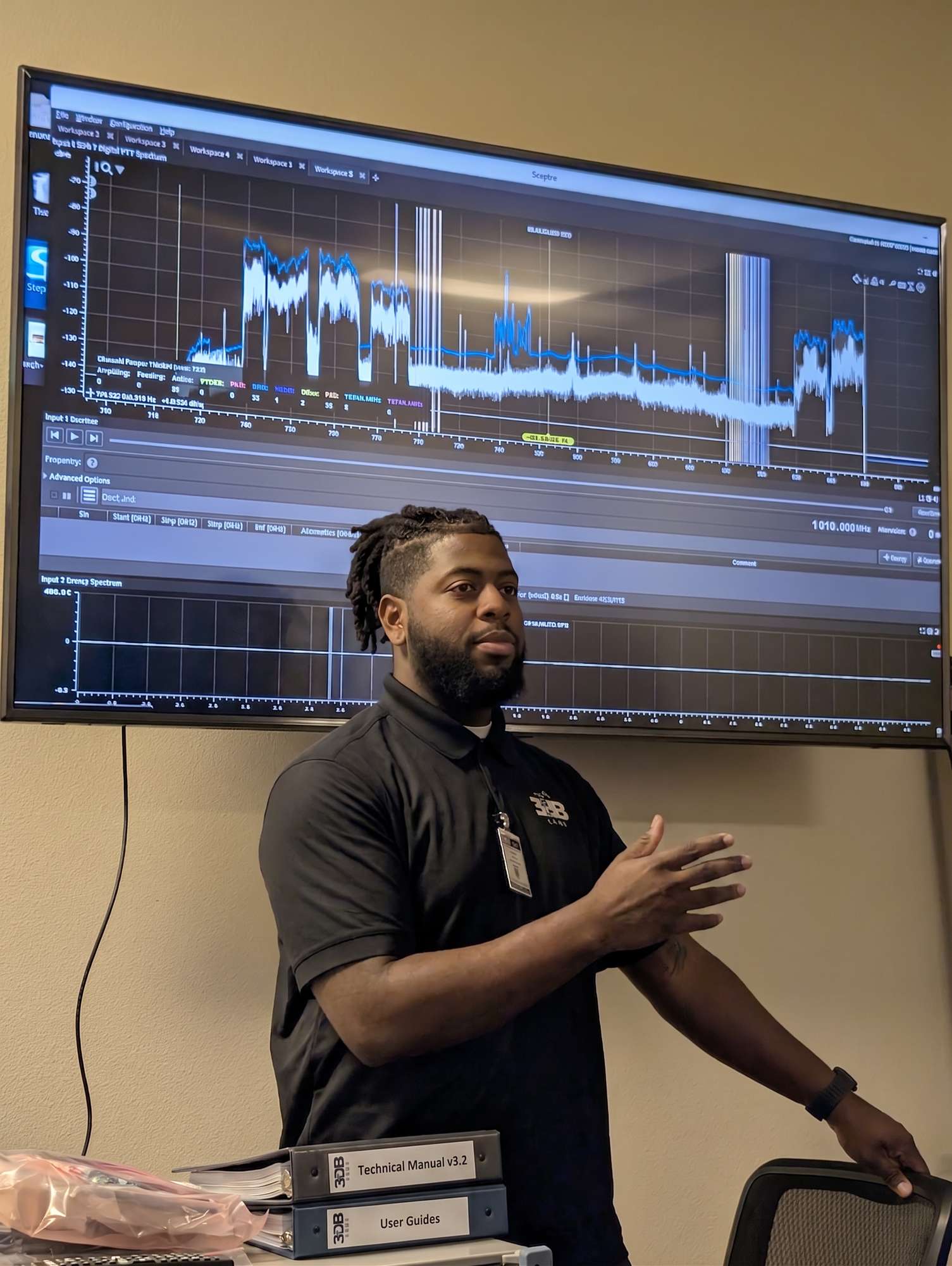 3dB Labs instructor teaching in front of a wall display running a live Sceptre spectrum workspace, with Sceptre technical manuals and user guides on the table